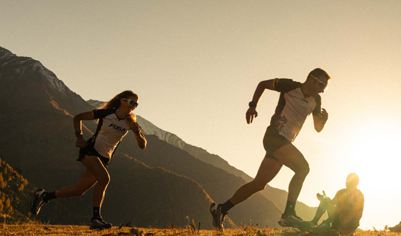 Two runners in trail gear sprint across a mountain ridge at golden hour, silhouetted against the sun.