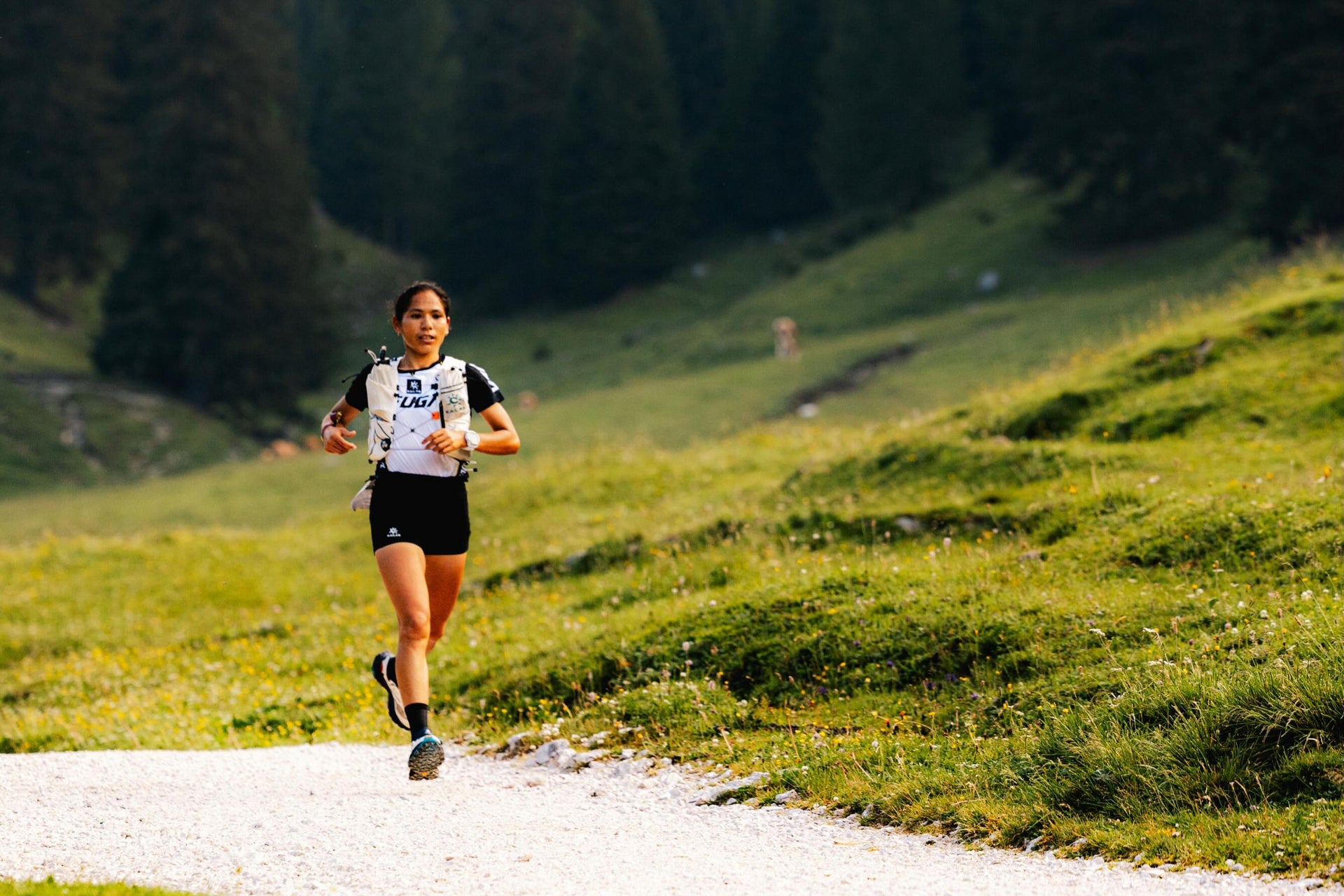 A female trail runner wearing a white and black bib top and black shorts is running on a gravel path in a lush green mountain valley with pine trees and distant snow-capped peaks.
